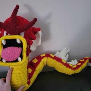 May include: A crocheted dragon toy with a red head, yellow and red body, and white teeth. The dragon has a pink tongue and is in a roaring pose.