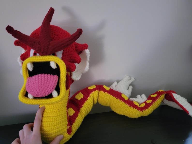 May include: A crocheted dragon toy with a red head, yellow and red body, and white teeth. The dragon has a pink tongue and is in a roaring pose.