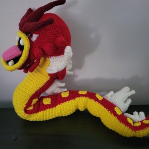May include: A crocheted red and yellow dragon with white accents. The dragon has a long, curled tail and a large, toothy mouth.