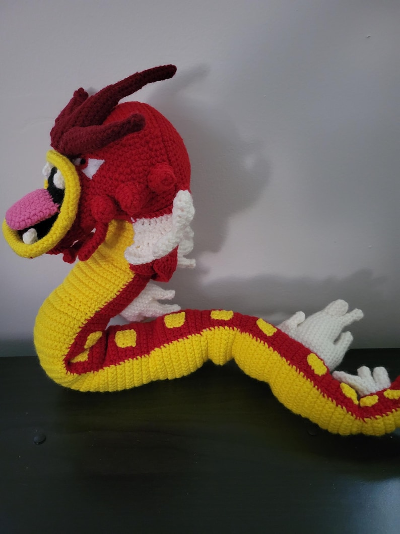 May include: A crocheted red and yellow dragon with white accents. The dragon has a long, curled tail and a large, toothy mouth.