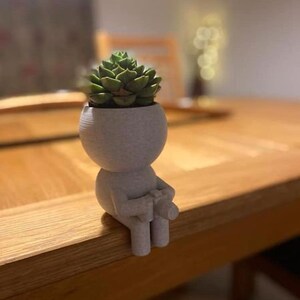 Puede incluir: Un pequeño macetero blanco impreso en 3D con forma de persona sentado en una mesa de madera. El macetero tiene una pequeña planta suculenta creciendo dentro.
