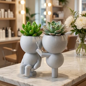 May include: A light gray speckled planter set featuring two stylized figures, one seated and one standing, with the standing figure holding small scissors. Each figure has a rounded head containing a green succulent plant.