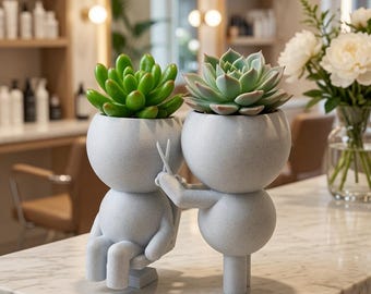 Può includere: Un set di vasi color grigio chiaro maculato con due figure stilizzate, una seduta e una in piedi, con la figura in piedi che tiene in mano delle piccole forbici. Ogni testa arrotondata contiene una pianta succulenta verde.