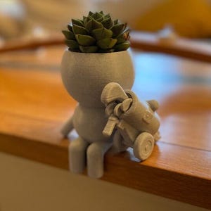 May include: A gray 3D printed planter shaped like a person sitting down with a succulent plant in the head. The planter has a small golf cart attached to the back.