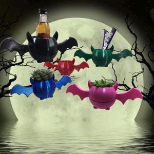 Può includere: Cinque vasi a forma di pipistrello appesi in diversi colori, tra cui nero, verde, rosso, blu e rosa. Ogni vaso ha una piccola pianta che cresce al suo interno. I vasi sono appesi a un ramo d'albero sullo sfondo di una luna piena.
