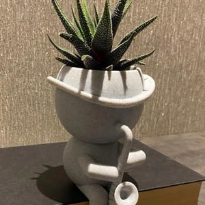 May include: A gray ceramic planter shaped like a person playing a saxophone. The planter has a wide brim hat and is holding a succulent plant.