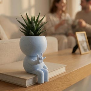 Op de afbeelding: Een lichtblauwe gespikkelde plantenbak in de vorm van een zittend figuur met een rond hoofd en lichaam. Het figuur heeft één arm omhoog in een handgebaar met de wijsvinger en pink uitgestoken. Een groene vetplant staat in het hoofd van het figuur.