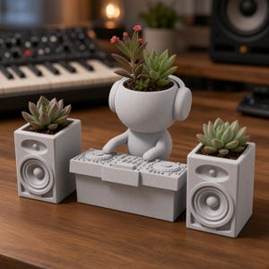 Peut inclure: Un ensemble de pots de fleurs gris clair mouchetés comprenant une figurine centrale avec un casque derrière une table de mixage DJ, entourée de deux pots rectangulaires en forme d'enceintes. Chaque pot contient une plante succulente verte.