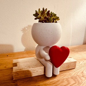 May include: A white speckled ceramic planter shaped like a person, holding a red heart. The planter contains a small succulent plant. The figure sits on a small wooden bench, a charming decorative piece.