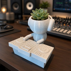 May include: A light blue, 3D-printed planter shaped like a character wearing headphones and operating a DJ controller. The character's head serves as a pot containing a green succulent plant, while the hands rest on the detailed mixing console.