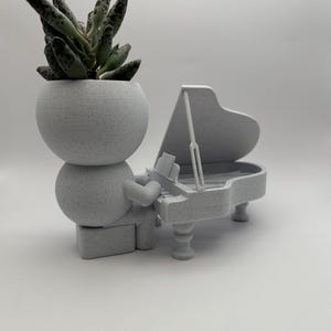 Könnte beinhalten: Ein weißer Keramik-Pflanztopf in Form einer Person, die ein weißes Keramik-Klavier spielt. Der Topf hat einen runden Kopf, aus dem eine grüne Sukkulente wächst.