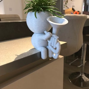 May include: A white ceramic planter shaped like a person playing a trumpet. The planter has a green succulent plant growing out of the top.