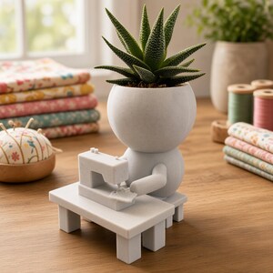 Può includere: Un vaso per piante grigio chiaro con finitura opaca a forma di figura stilizzata seduta a un tavolo da cucito. La testa della figura funge da vaso contenente una pianta succulenta verde e appuntita. L'oggetto è realizzato in materiale solido.