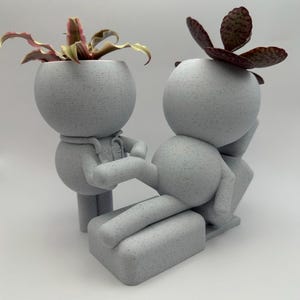 May include: A white ceramic planter shaped like a person sitting in a chair, with a green plant in the head of the person in the chair and a pink plant in the head of the person standing.