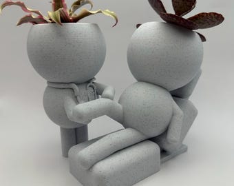 Può includere: Un vaso di fiori in ceramica bianca a forma di persona seduta su una sedia, con una pianta verde nella testa della persona seduta e una pianta rosa nella testa della persona in piedi.