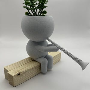 Puede incluir: Maceta de cerámica blanca con forma de persona tocando un clarinete. La maceta está sentada en una repisa de madera y tiene una pequeña planta verde creciendo dentro.