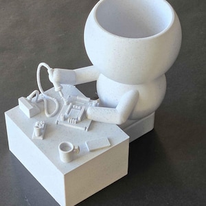 Op de afbeelding: Witte keramische figuur van een persoon die aan een bureau zit met een soldeerbout en een printplaat. De figuur heeft een groot hoofd en een klein lichaam. Het bureau heeft een kopje en een telefoon erop.