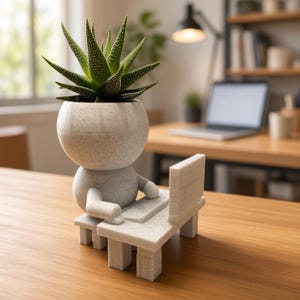 May include: A light gray, speckled, 3D-printed planter shaped like a small figure sitting at a desk with a laptop. The figure's head serves as a pot containing a green, spiky succulent plant. The entire structure is made of a matte, textured material.