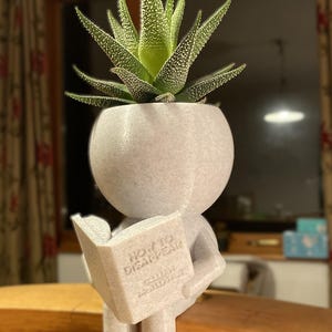 Op de afbeelding: Een witte 3D-geprinte plantenpot in de vorm van een persoon die een boek leest met de titel "How to Disappear" van Gillian Holloway. De pot bevat een groene succulent.