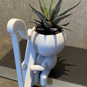 Könnte beinhalten: Ein weißer 3D-gedruckter Pflanztopf in Form einer Person, die eine Pflanze hält. Die Pflanze ist eine Sukkulente mit grünen Blättern und weißen Flecken. Der Topf steht auf einem schwarzen Buch.