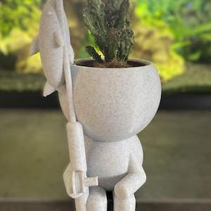 Może przedstawiać: Biała ceramiczna doniczka w kształcie siedzącej osoby trzymającej wędkę z delfinem na końcu. Doniczka ma chropowatą fakturę i zawiera małą zieloną roślinę kaktusową.