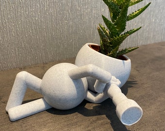 Può includere: Un vaso di ceramica bianco a forma di persona che tiene una pianta. La pianta è una succulenta verde con foglie appuntite. Il vaso è sdraiato su un lato.