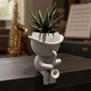 May include: A matte light gray planter shaped like a stylized figure wearing a hat and playing a saxophone. The figure is seated with legs dangling over the edge. A green succulent plant with pointed, textured leaves grows from the hat.
