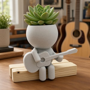 May include: A light gray speckled planter shaped like a stylized figure sitting on a light wood block. The figure has a round head containing a green succulent plant and holds a matching light gray guitar. The design is smooth and minimalist.