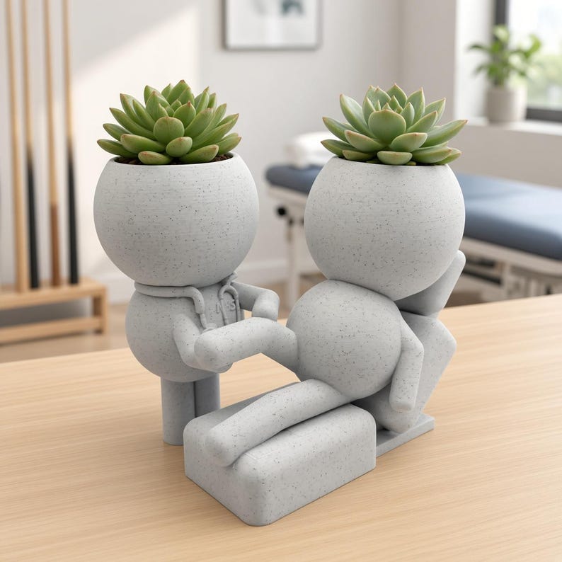 Pu&ograve; includere: Un set di vasi grigio chiaro maculato con due figure stilizzate. Una figura &egrave; in piedi mentre l'altra &egrave; sdraiata su una base rettangolare. Ogni testa rotonda funge da vaso, con una pianta succulenta verde piantata nell'apertura superiore.