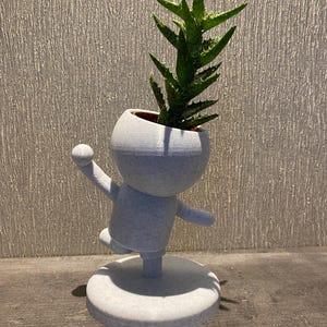 Puede incluir: Macetero de cerámica blanca con forma de persona con una planta suculenta verde que crece fuera de su cabeza. El macetero está de pie sobre una base redonda.