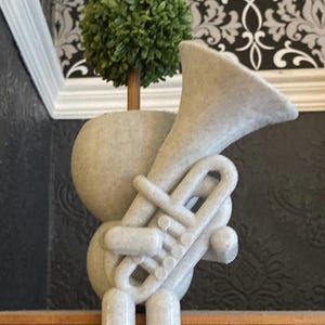 Puede incluir: Una escultura de cerámica blanca de una persona sosteniendo una trompeta. La persona está de pie sobre una superficie de madera. Una pequeña planta verde en maceta está detrás de la escultura.