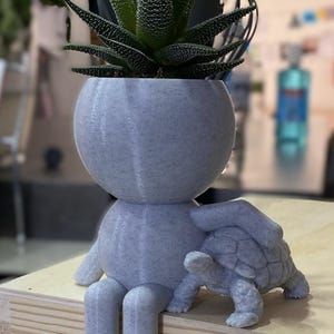 May include: A gray, 3D printed planter shaped like a person sitting on a wooden surface. The planter has a round head and is holding a small, gray, 3D printed turtle. A green succulent plant is growing in the planter.