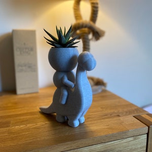 May include: A gray ceramic planter shaped like a person riding a dinosaur. The planter has a small succulent plant growing inside.