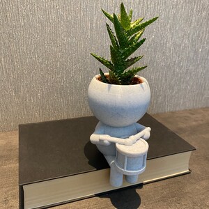 May include: A white ceramic planter shaped like a person playing a drum, with a small green succulent plant growing inside. The planter is sitting on a black book.