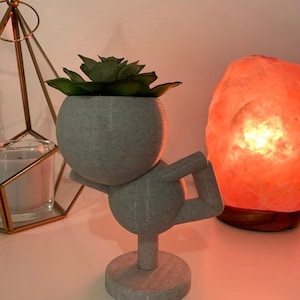 Op de afbeelding: Een grijze keramische plantenpot in de vorm van een persoon in een yogapose, die een kleine succulent plant vasthoudt. De pot heeft een getextureerd oppervlak en staat op een wit oppervlak.