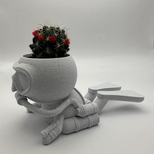 May include: White ceramic planter shaped like a scuba diver with a cactus plant inside. The planter has a round head with a diving mask, a body with a tank, and two flippers.