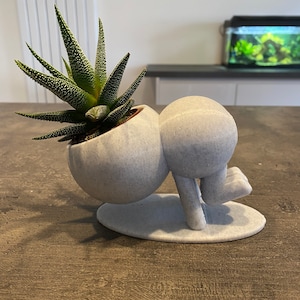 May include: White ceramic planter shaped like a person doing a handstand, with a green succulent plant growing out of the top.