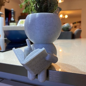 Può includere: Un vaso maculato blu chiaro a forma di figura che legge un libro, con un cactus. La figura è seduta sul bordo di un bancone. Il libro ha del testo sulle pagine. Il cactus è verde.