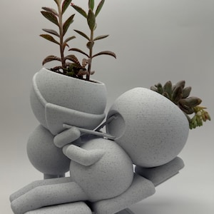 Puede incluir: Una maceta gris claro, impresa en 3D, con la forma de una persona reclinada en un banco, sosteniendo una maceta pequeña. La maceta contiene dos suculentas con hojas verdes y rojas. El diseño es moderno y minimalista.