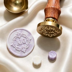 May include: A brass wax seal stamp with a wooden handle, a round lavender wax seal featuring the letters A|G inside an ornate border, a small white wax bead, a small lavender wax bead, and a brass melting spoon for wax sealing projects.