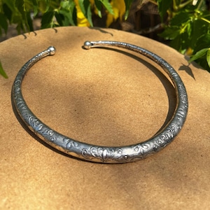 Puede incluir: Un collar gargantilla plateado, abierto, con un patrón floral en relieve. El collar tiene forma redondeada y se muestra sobre una superficie marrón, con follaje verde en el fondo.