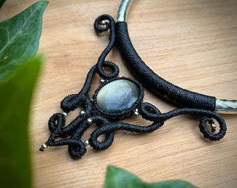 May include: A handcrafted black macrame necklace with a silver-toned metal collar. The pendant features a large, oval-shaped, iridescent stone. The necklace is displayed on a wooden surface, with green foliage in the background.