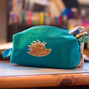 May include: A teal blue zippered pencil case with a brown and gold hedgehog design. The case is filled with pens and pencils.
