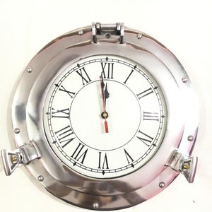 May include: A silver round clock with Roman numerals and a white face. The clock is in a porthole style with a silver metal frame and a glass cover. The clock is set to 12 o'clock.