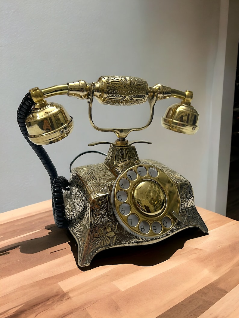 May include: A gold rotary phone with an ornate design and a black coiled cord. The phone has a round dial with numbers 1 through 0.