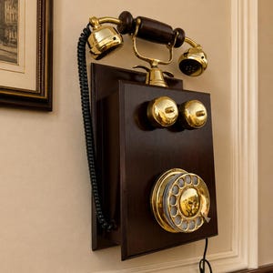 Puede incluir: Un tel&eacute;fono antiguo montado en la pared con un gabinete de madera oscura y detalles de lat&oacute;n. El tel&eacute;fono tiene un dial giratorio, dos campanas de lat&oacute;n y un cable de auricular negro en espiral. El dise&ntilde;o es reminiscente de la tecnolog&iacute;a de principios del siglo XX.