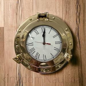 May include: A gold-toned round clock with Roman numeral markings on a white face. The clock is in a brass porthole-style frame with a woodgrain background.