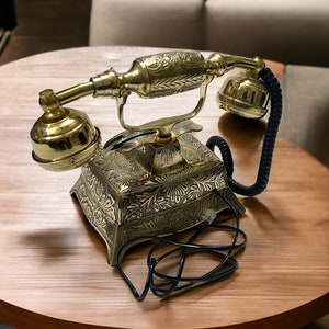 May include: A gold-tone rotary phone with intricate floral designs on the base and receiver. The phone has a black cord and is sitting on a wooden table.