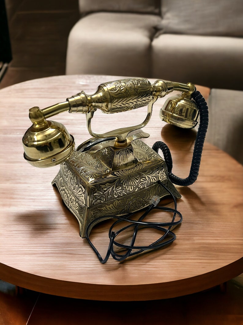 May include: A gold-tone rotary phone with intricate floral designs on the base and receiver. The phone has a black cord and is sitting on a wooden table.