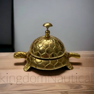 May include: A gold-colored turtle-shaped service bell with a hexagonal pattern on the top and bottom. The bell is made of metal and has a small knob on top.
